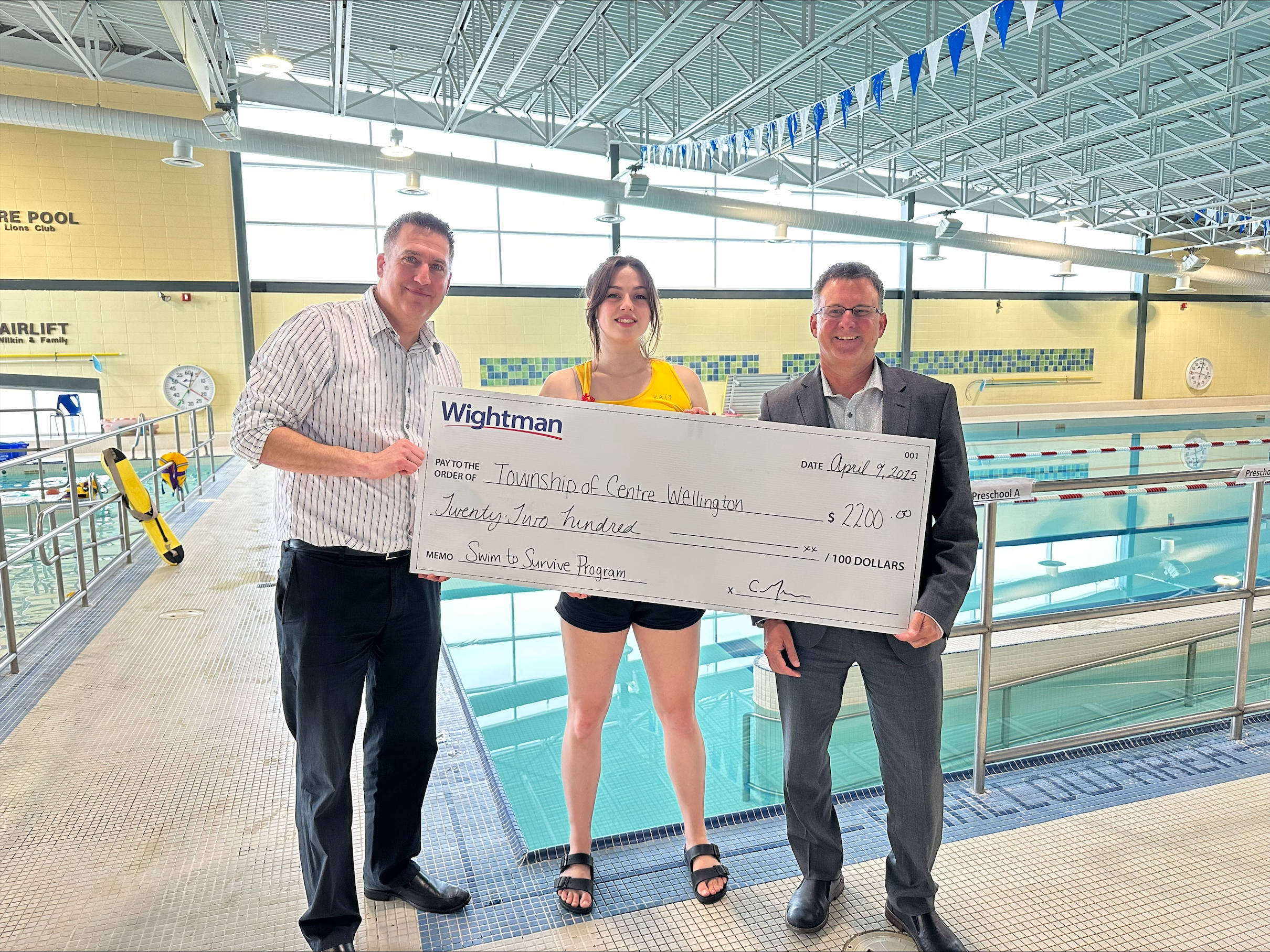 Three individuals holding a large cheque standing beside the Fergus Public Pool.